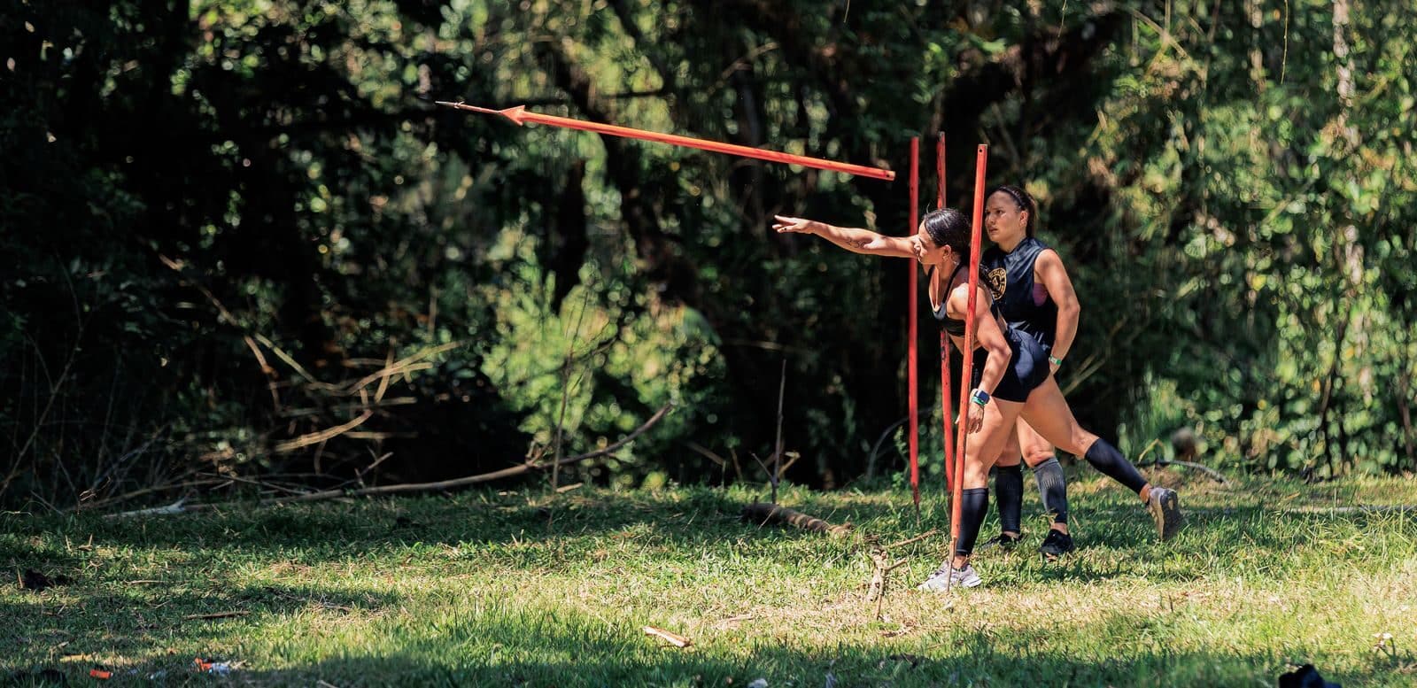 Two women practicing javelin throw outdoors in a lush forest setting in Colombia.